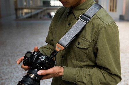 Male model wearing a dark grey camera strap with rust coloured accents, looking at the rear LCD screen of a Nikon DSLR camera.